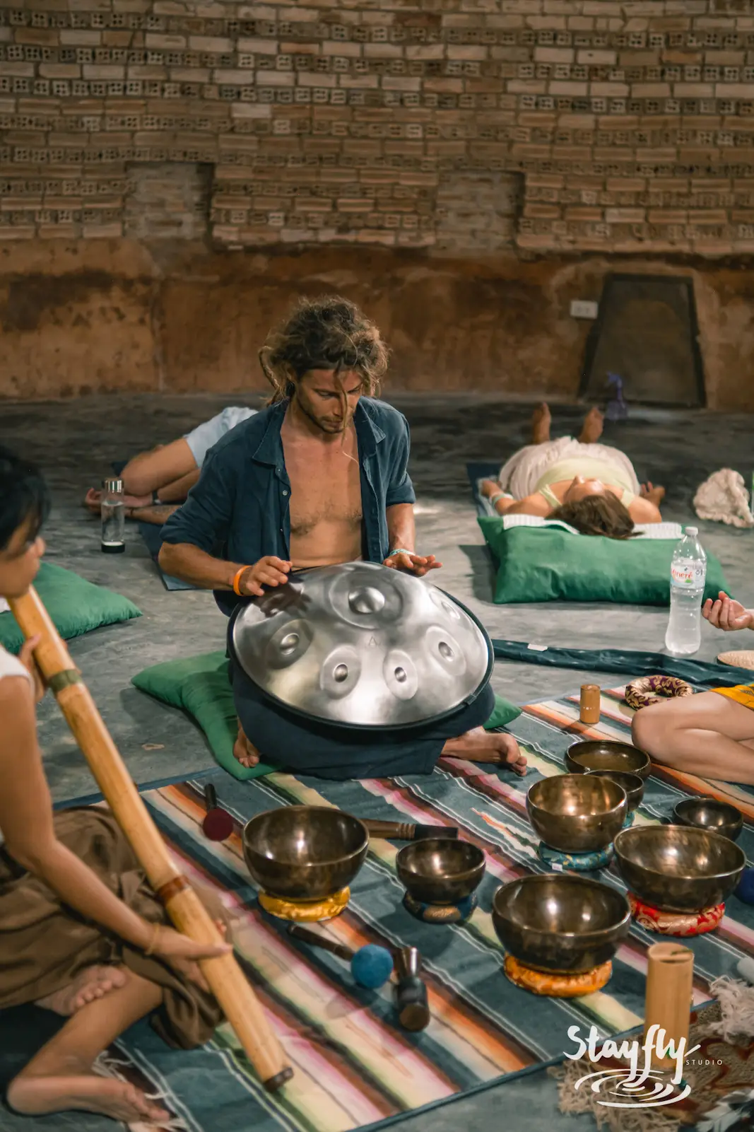 A handpan played with both hands on the ground, singing bowls waiting within reach.