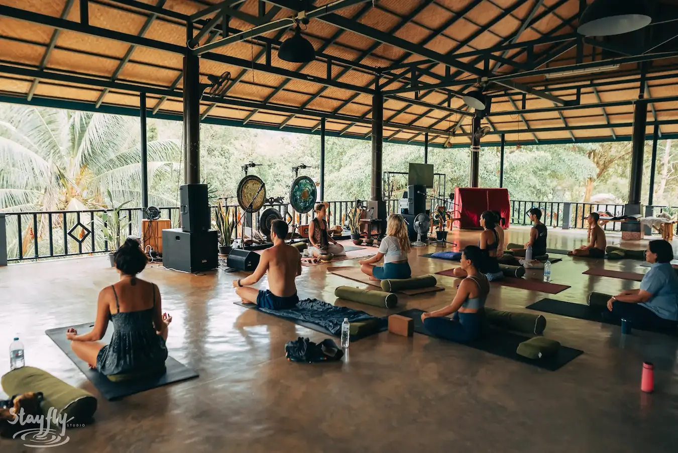 A wide view of the pavilion, participants seated on mats facing gongs hung along the railing.