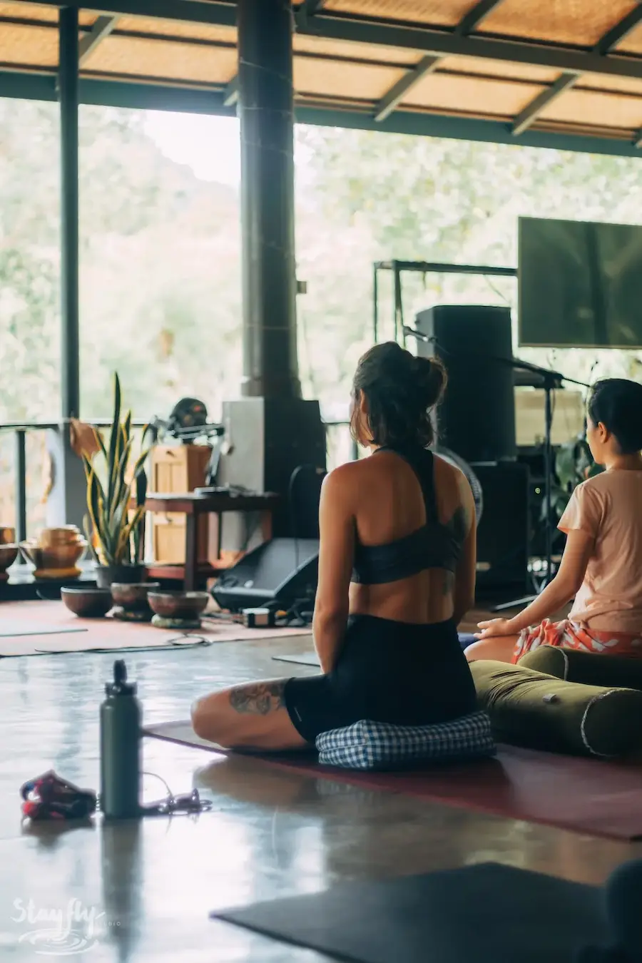 A woman sits cross legged on her mat in the pavilion, gongs hanging behind her and trees beyond.
