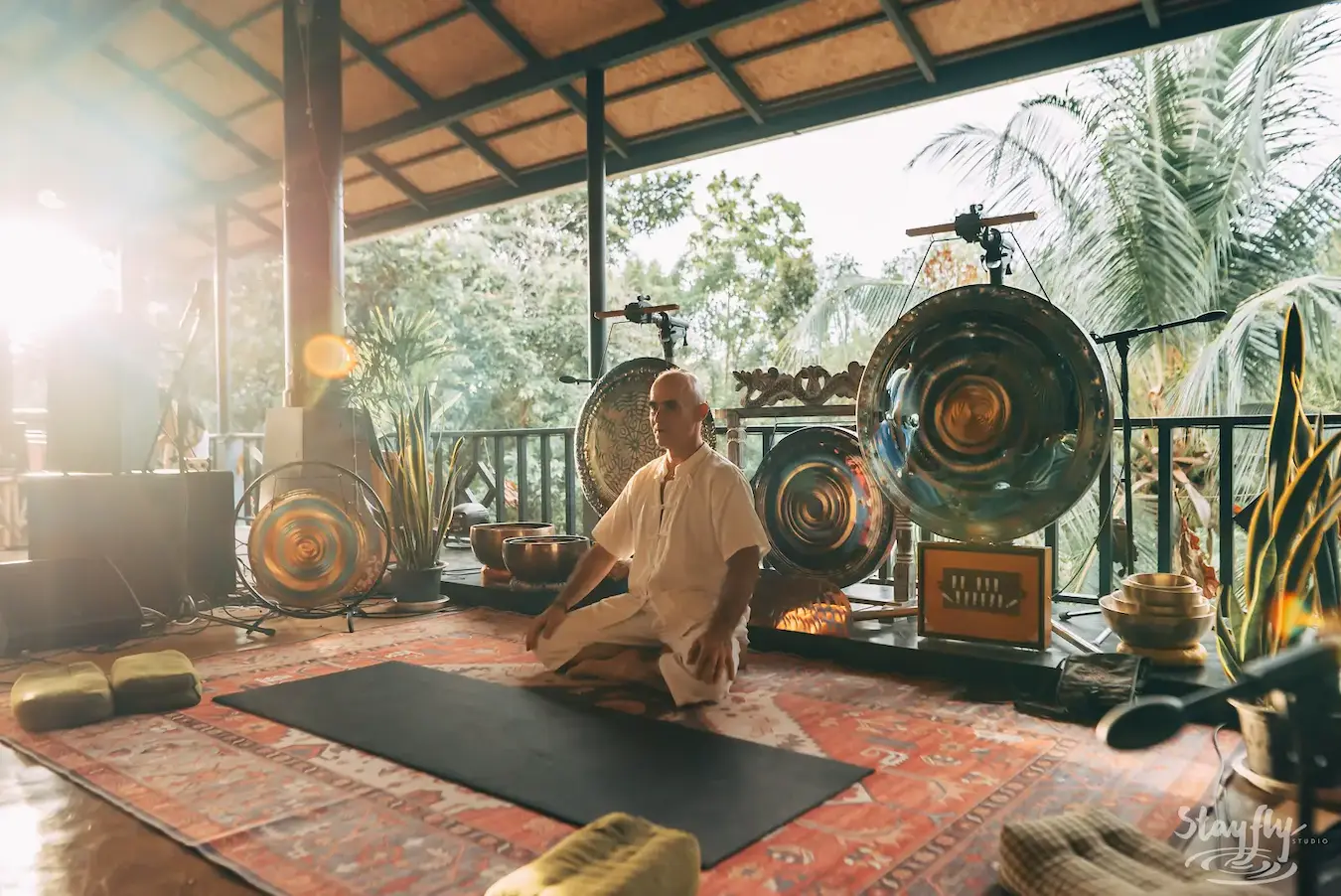 A woman in white sits in meditation on a rug with gongs and singing bowls around her.