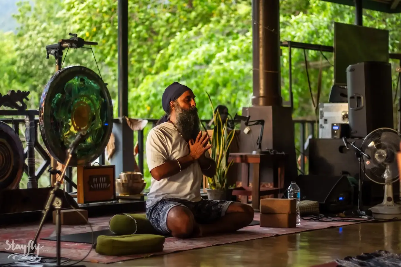 A bearded practitioner sits in the pavilion beside a gong and a singing bowl, hands together.
