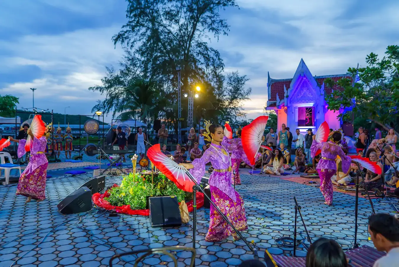 Dancers with red and white fans perform on the pier at dusk beside an illuminated stage.