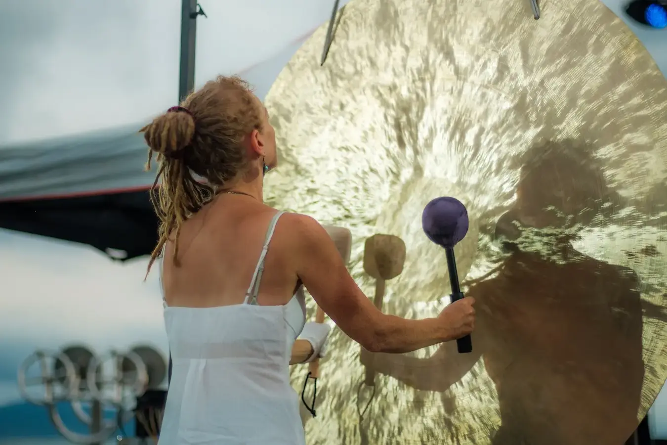 A woman in white plays a large gong with a mallet, the metal filling the frame.
