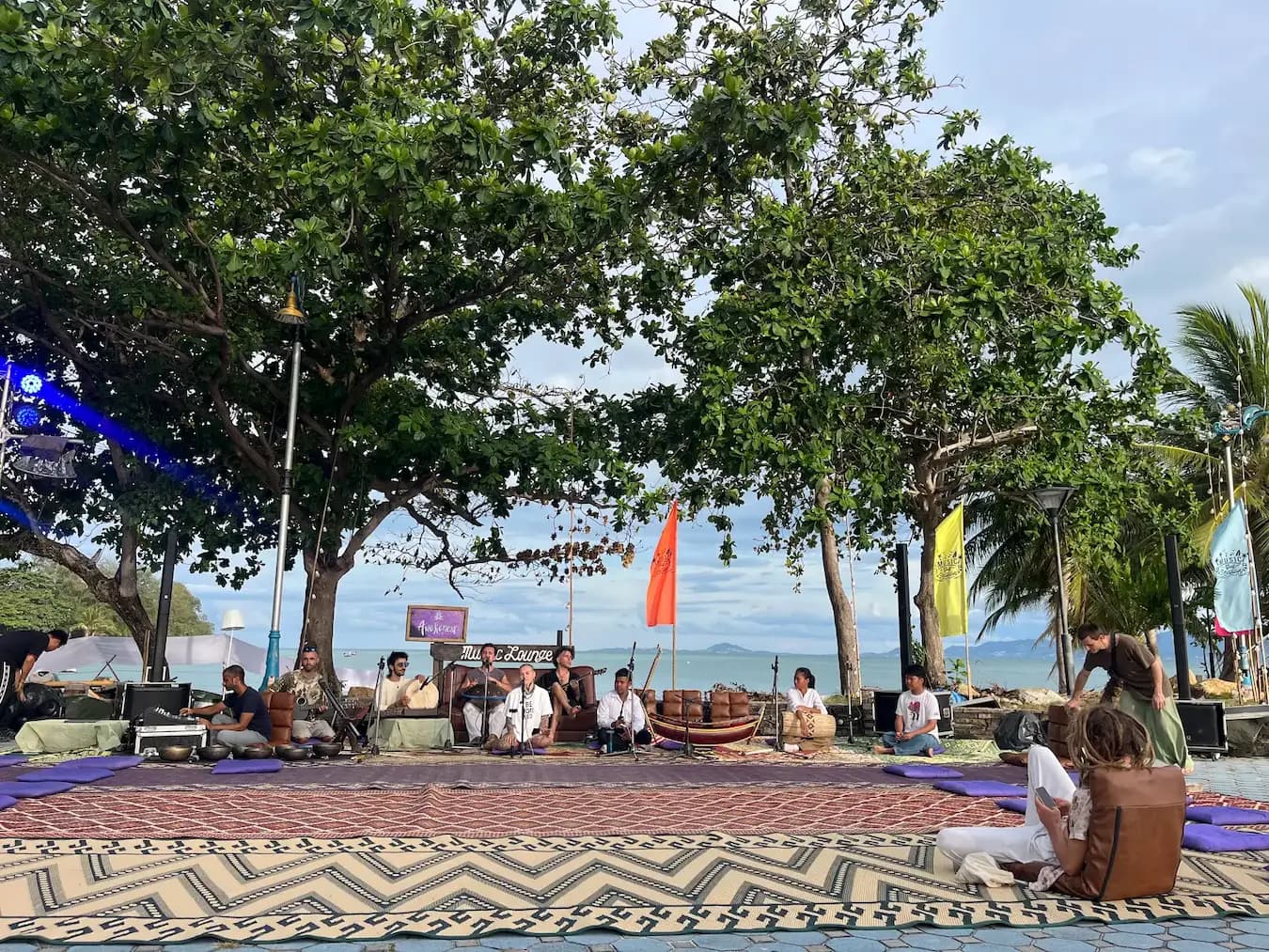 A large tree with festoon lights and orange flags in front of the sea, people seated on mats beneath it.