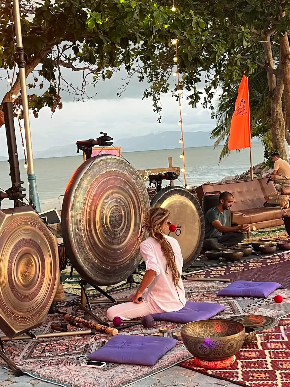 Gongs on stands and a frame drum laid out on rugs beneath the trees by the sea.