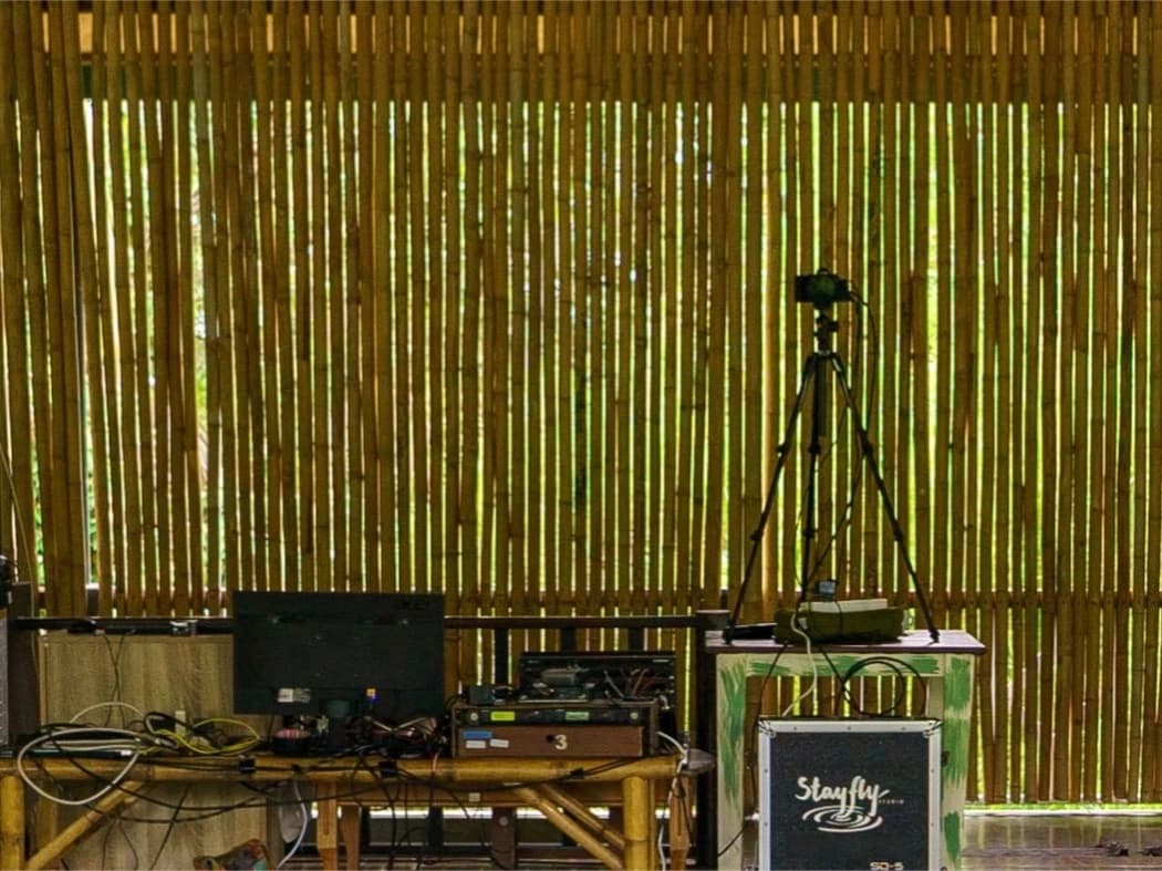 A control position against a bamboo wall: a monitor, an audio interface, a camera on a tripod and a Stayfly Studio flight case.