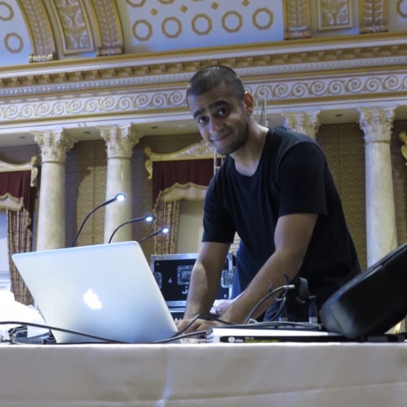 Adisorn Kukarja at a laptop and mixing desk in an ornate hall with a gilded ceiling and columns.