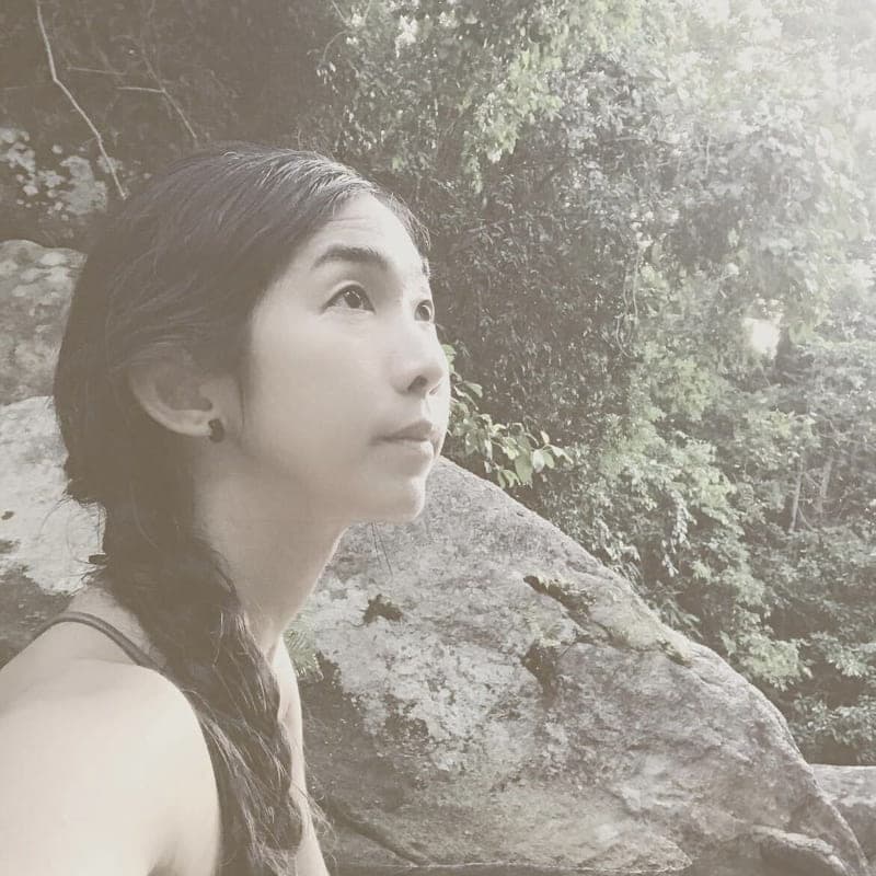 Khane Chompreeda looking upward beside a large boulder, with forest behind her.