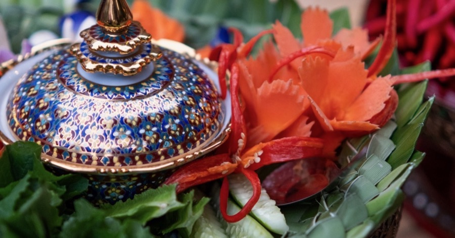 Carved carrot flowers, red chillies, cucumber and greens arranged around a painted benjarong bowl on woven banana leaf.